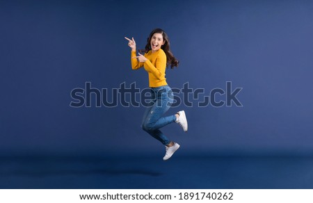 Happy young Asian woman in colorful yellow color casual clothes jumping and presenting to copy space on blue color background, lifestyle and leisure, travel and tourist, travel and vacation concept