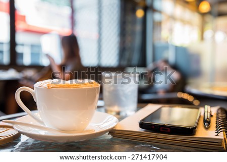 Cup of cappuccino with note book and pen, blur coffee shop background