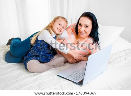 happy blonde hair daughter with her brunette mother sitting on bed working together on computer