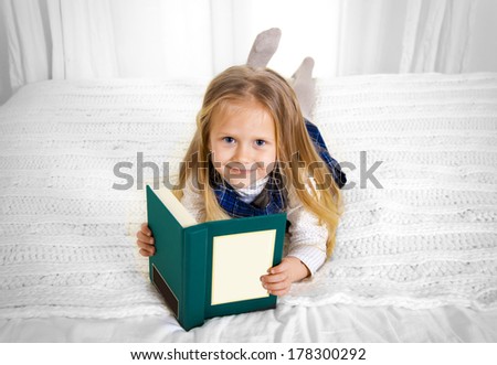 happy cute blonde haired school girl wearing a school uniform reading a book smiling at the camera lying on the bed