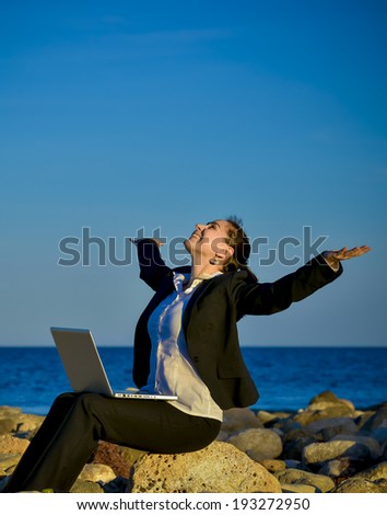 happy attractive business woman working on a laptop sitting in front of the sea on some rocks at the beach looking up to blue sky with opened arms thinking about freedom