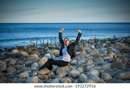 happy attractive business woman wearing suit working on computer sitting on some rocks at the beach looking up to sky thinking about freedom