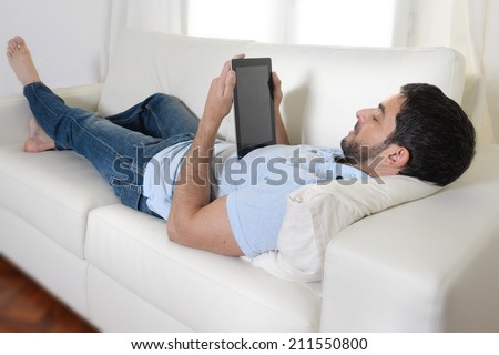 young happy attractive man using digital pad or tablet lying relaxed on couch at home connected to internet reading , watching the screen and smiling pensive wearing casual blue shirt and jeans