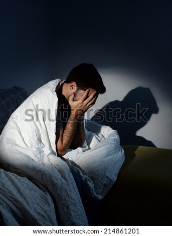 young sick looking man sitting on couch at home scary desperate looking, suffering insomnia, depression, nightmares, emotional crisis or mental disorder with dim light and deep dark shadow on the wall