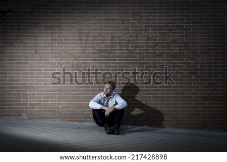 Young business man who lost job abandoned lost in depression sitting on ground street corner against brick wall suffering emotional pain, thinking and crying alone