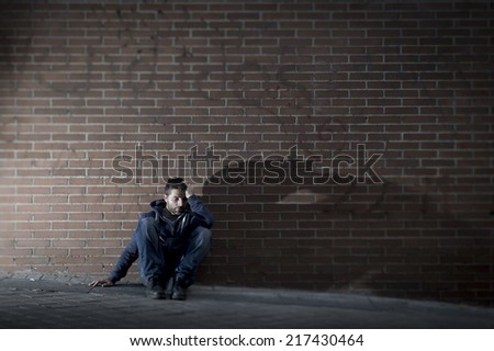 Young desperate man who lost job abandoned and lost in depression sitting on ground street corner against brick wall suffering emotional pain, crying alone in grunge lighting