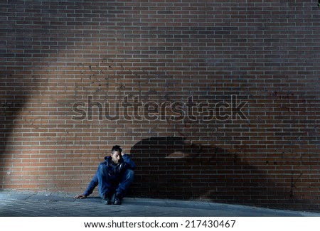 Young desperate man who lost job abandoned and lost in depression sitting on ground street corner against brick wall suffering emotional pain, crying alone in grunge lighting