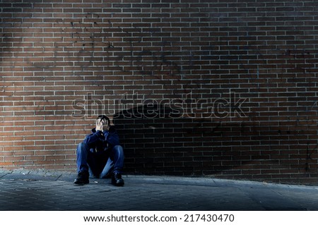 Young desperate man who lost job abandoned and lost in depression sitting on ground street corner against brick wall suffering emotional pain, crying alone in grunge lighting