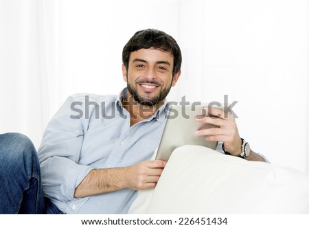 Young attractive hispanic man happy at home lying on couch smiling using digital tablet or pad looking relaxed at living room enjoying surfing internet watching online movie