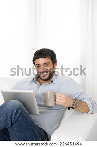 Young attractive hispanic man at home sitting on white couch using digital tablet or pad looking relaxed with coffee at living room enjoying surfing internet watching online movie