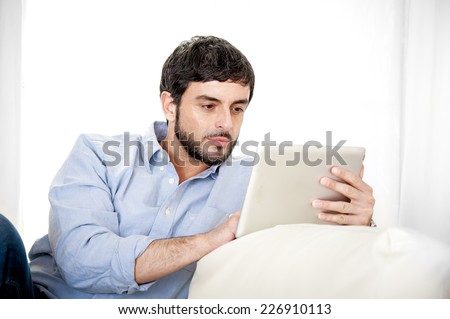 Young attractive Hispanic man at home sitting on white couch using digital tablet or pad looking relaxed at living room enjoying surfing internet and working online