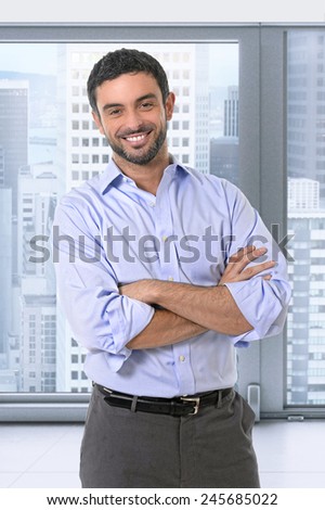 attractive businessman standing in corporate portrait smiling with folded arms in smart casual shirt and suit trousers in front of office window business district view in work and success concept