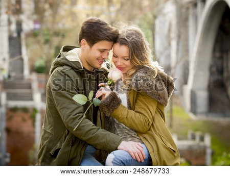 candid portrait of beautiful European couple with rose in love kissing on street  celebrating Valentines day with passion sitting on city park in winter urban background