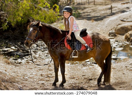 sweet beautiful young girl 7 or 8 years old riding pony horse hugging and smiling happy wearing safety jockey helmet posing outdoors on countryside in summer holiday