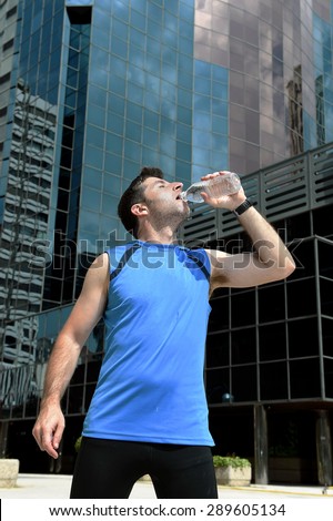 young sport man drinking water bottle after running training session in business district with office buildings and blue sky background in healthy lifestyle and fitness concept