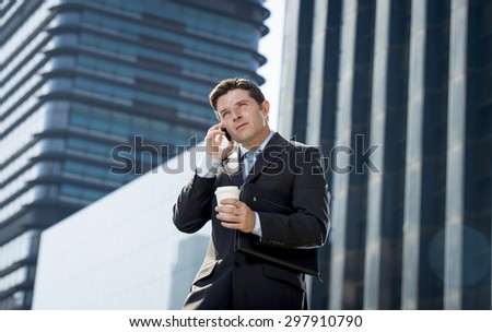 young attractive businessman in suit and necktie talking on mobile smart phone holding take away coffee standing outdoors in exterior office buildings on business district in success concept