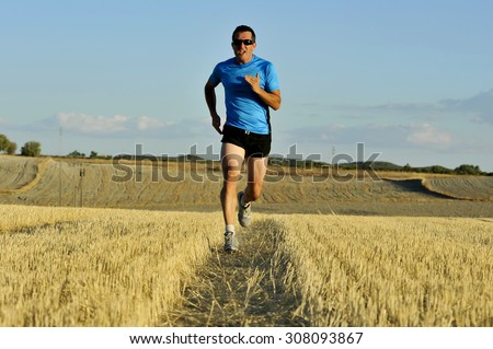 young sport man with sunglasses running outdoors on straw field ground in frontal perspective towards camera in healthy lifestyle and summer training concept