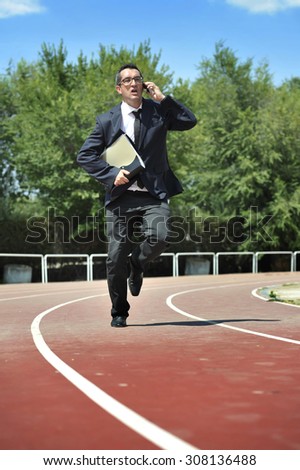 businessman in suit and necktie carrying folder portfolio and files running in stress on athletic track talking on mobile phone looking tired and stressed in work urgent problems concept