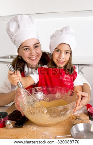 happy mother baking with little daughter in apron and cook hat working with flour , bowl and spoon mixing dough for muffins teaching the kid cooking and having fun together