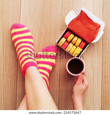Comfort Concept - Woman drinking hot cocoa and eating delicious macaroon. Close-up of female legs in bright colored warm socks with a retro vintage instagram filter.