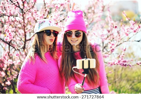 Closeup fashion lifestyle portrait of two young pretty beautiful friends in the lush spring garden on a sunny day. Cheerful friends, having fun together, joy and making selfie with self monopod.