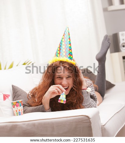 Red Headed very cute girl smiling on her sofa in her living room having some fun in a party with a carton cone hat, a drink and party-props