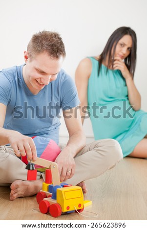 pregnant woman worried about readiness of his husband.  He plays with wooden toys. They wear blue and cyan clothes