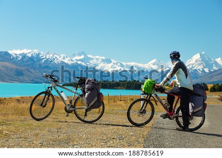 cyclist stands on the winding asphalt mountain road near Lake Pukaki view from Glentanner Park Centre near Mount Cook, on a background of blue sky with clouds, snowy Southern Alps.