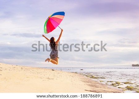 young woman jumping high with joy on the beach in summer with a colorful umbrella on a background of blue sky with clouds