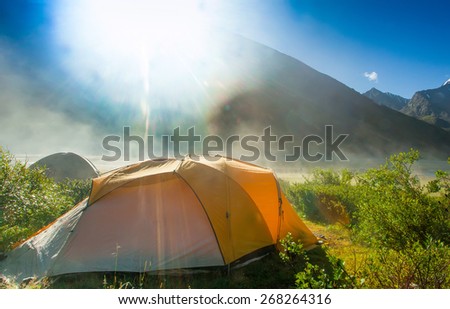 Tourist tent in forest camp in the meadow, in the background of high snow-capped mountains. misty morning and bright sunshine after the rain