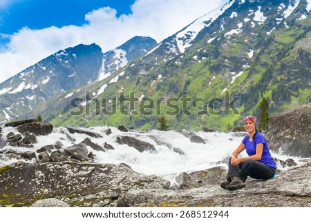 Healthy hiker girl in nature hike. Beautiful young woman hiking happy sits on a rock near a small waterfall. Background beautiful high mountains with snow-capped peaks, blue sky.