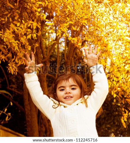 Photo of sweet small girl jumping in autumn forest, lovely female child playing on backyard, adorable little kid in warm sweater with raised hands having fun in fall woods, happy childhood concept