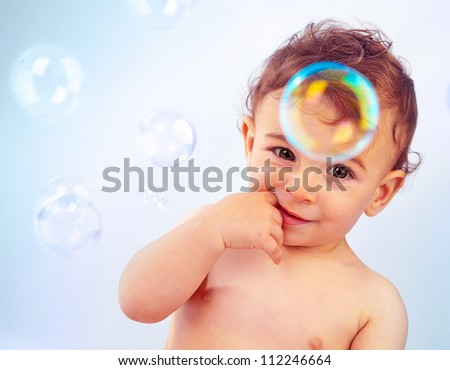 Closeup portrait of naked nice toddler isolated on blue background, cute happy baby boy bathe, adorable child sitting with finger in mouth, infant play with soap-bubbles, healthy childhood