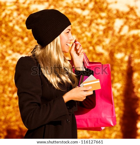 Photo of cute blond girl with pink shopping bag, side view of pretty woman holding paper present bag, beautiful happy shopper over autumn yellow foliage background, seasons sales