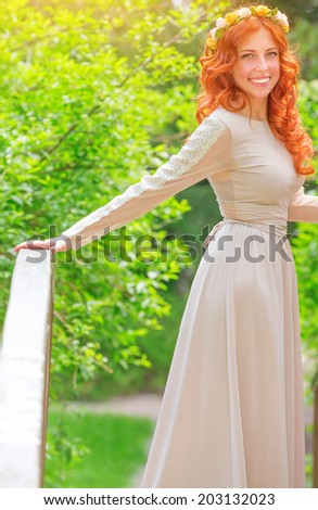 Beautiful happy bride wearing long beige dress and flower wreath in red hair, standing on little bridge in fresh summer park, happiness and love concept