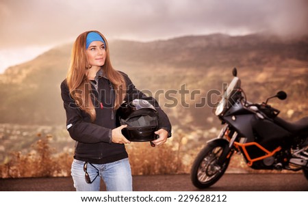 Beautiful biker girl standing on the road with helmet in hands near motorbike, traveling in overcast weather along high mountains, active lifestyle concept