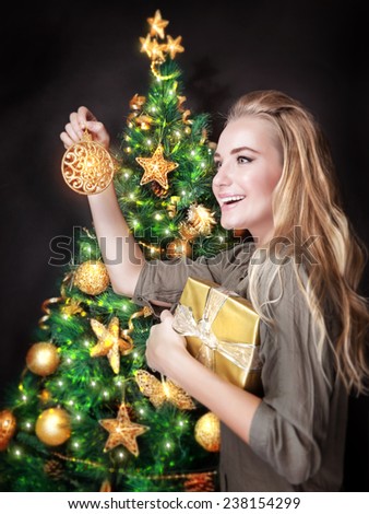 Portrait of cute blond female decorating beautiful Christmas tree over dark background, holding in hands little golden gift box, happy holidays concept