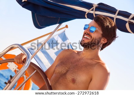 Portrait of cheerful captain behind wheel of luxury sailboat, sexy man wearing stylish sunglasses and enjoy interesting water trip, happy summer vacation concept
