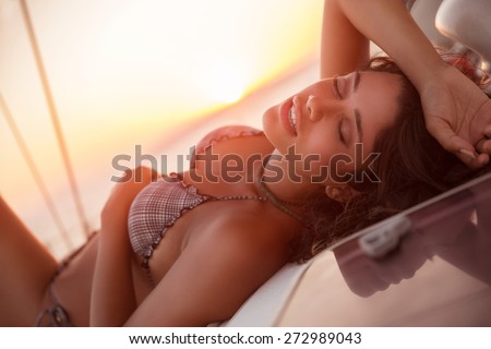 Gorgeous female with closed eyes of pleasure lying down on sailboat, tanning in mild sunset light, spending leisure time in the sea, luxury summer vacation concept