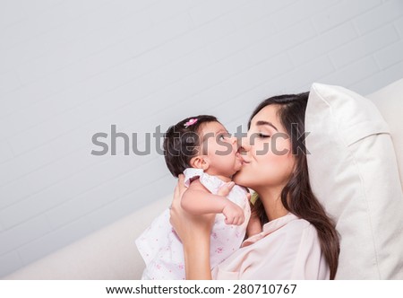Happy mother kissing baby, cheerful joyful mom lying down with her adorable daughter on the couch at home, loving young family