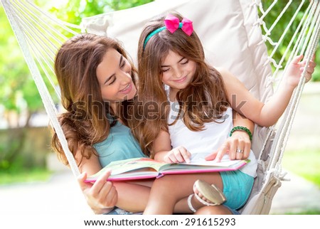 Portrait of beautiful mother with cute little daughter sitting on backyard in hammock and reading funny story, happy family life