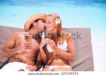 Loving couple on the beach, happy young family relaxing on sunbeds and refreshing cold tasty ice cream, handsome guy kissing his cute wife, happy summer holidays