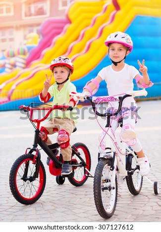 Happy friends on the bicycles, brother with sister having fun in amusement park, gesturing peace by hands, playing game outdoors, enjoying friendship and summer holidays