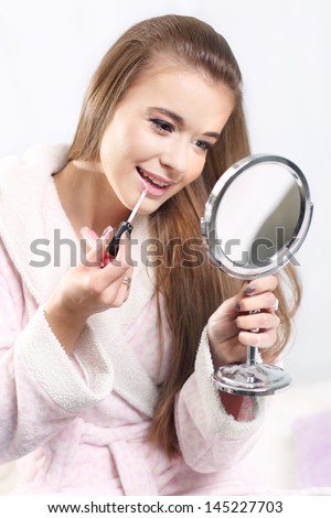 Young long-haired blonde girl in a bathrobe , holding lip gloss and mirror