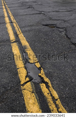 Pothole and cracked road with yellow dividing line  in need of repair.