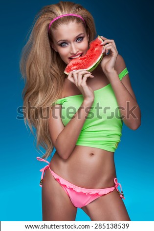 Sexy beautiful blonde happy woman eating fresh healthy watermelon, looking at camera. Girl posing on blue background. Summer photo. Young attractive lady with blue eyes.