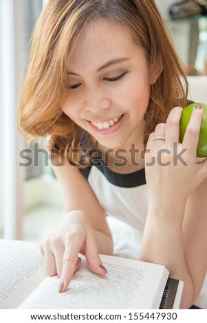 A woman is reading her book and holding a green apple in her room.