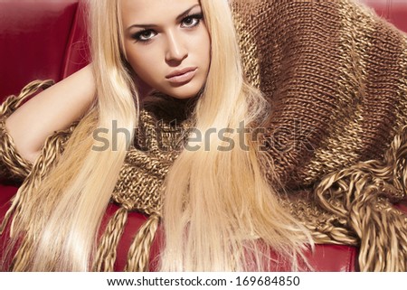 Beautiful Blond Woman on Red Leather Sofa. Beauty Fashion Girl in Poncho. Long Hair