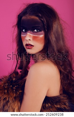young woman with tribal makeup dressed in leather and fur looking wild on pink background