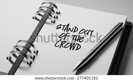 Closeup of a personal agenda setting an important date writing with pen. The words Stand out of the crowd written on a white notebook to remind you an important appointment.